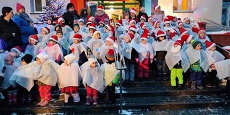 Powiększ grafikę: Przedszkolaki śpiewają kolędy na schodach wejściowych do przedszkola ubrane w białe pelerynki i mikolajowe czapeczki.